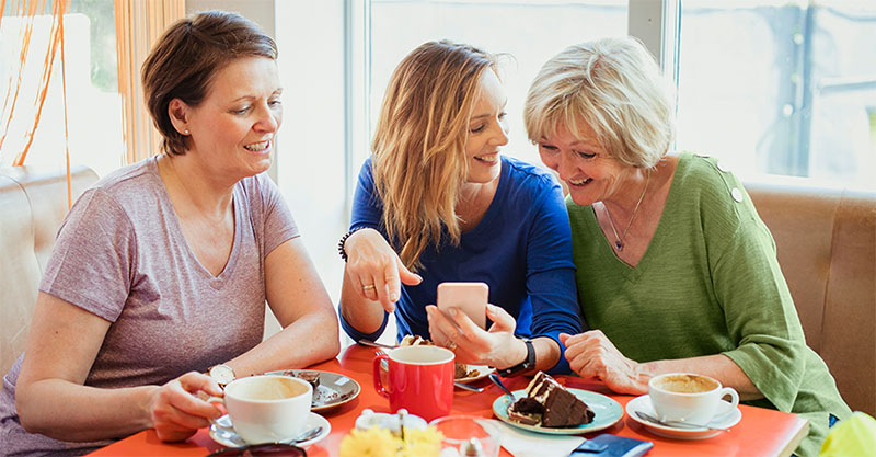 Three women at a table having coffee and dessert, gathered around looking at a mobile phone