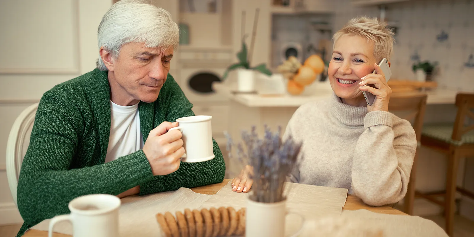 couple sitting at dining table, man drinking from mug and woman smiling while on phone